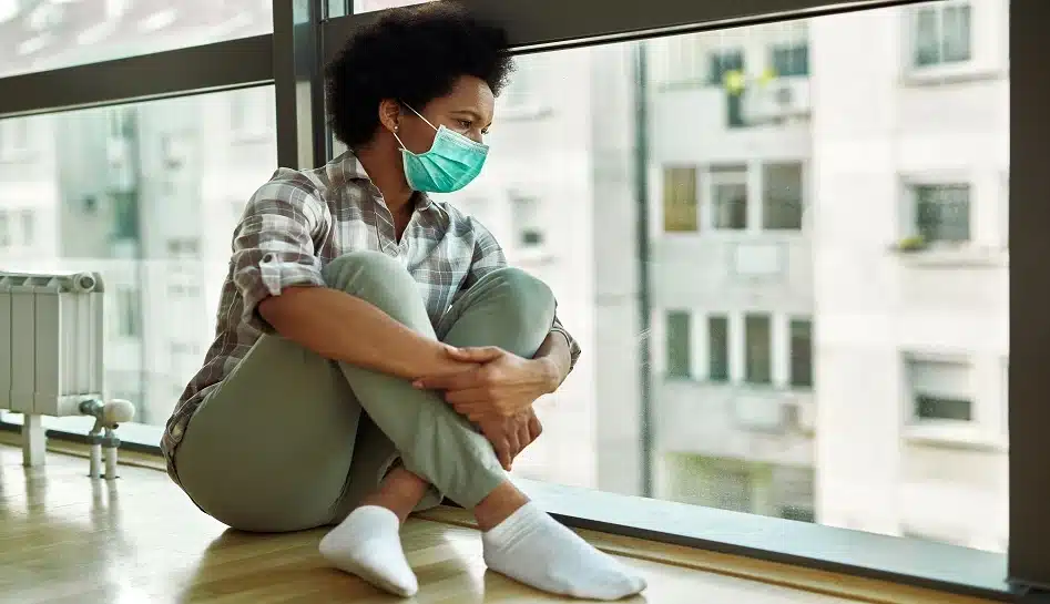 pensive-black-woman-with-face-mask-looking-through-window-while-sitting-floor-home